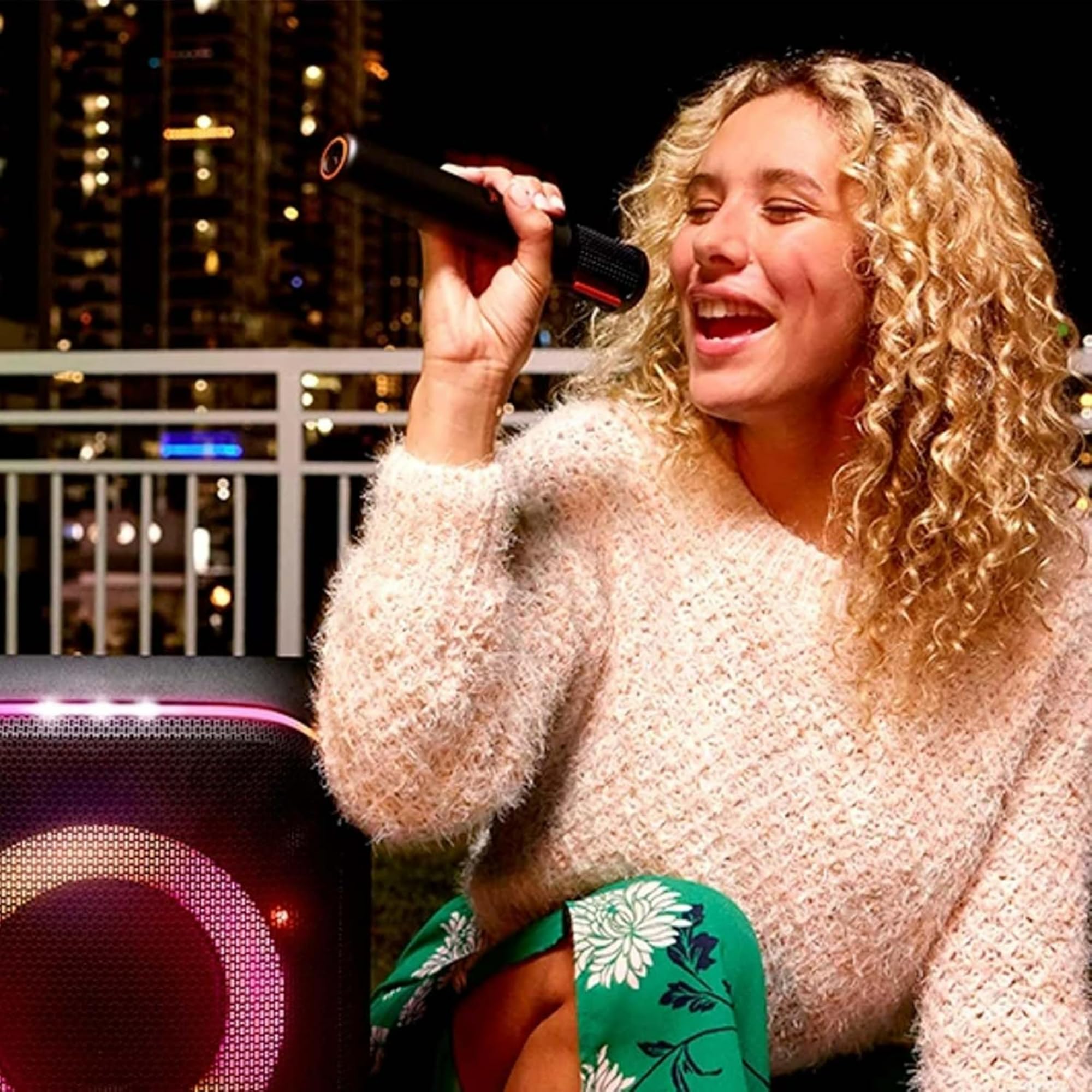 Woman singing into a mic beside a JBL PartyBox speaker with purple light rings