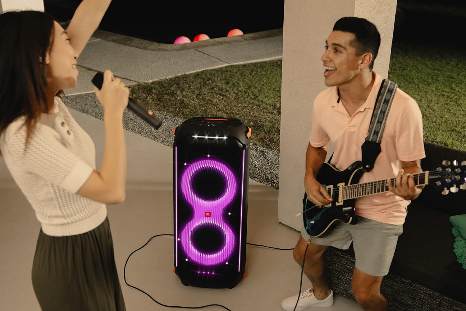 Woman sings while a man plays guitar beside a JBL PartyBox speaker with purple lights
